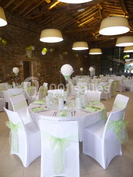 Salle de réception décorée avec des tables rondes, nappes blanches, chaises ornées de nœuds verts et centres de table…