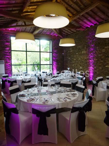 Salle de réception avec tables rondes dressées, chaises blanches ornées de nœuds noirs et éclairage violet.
