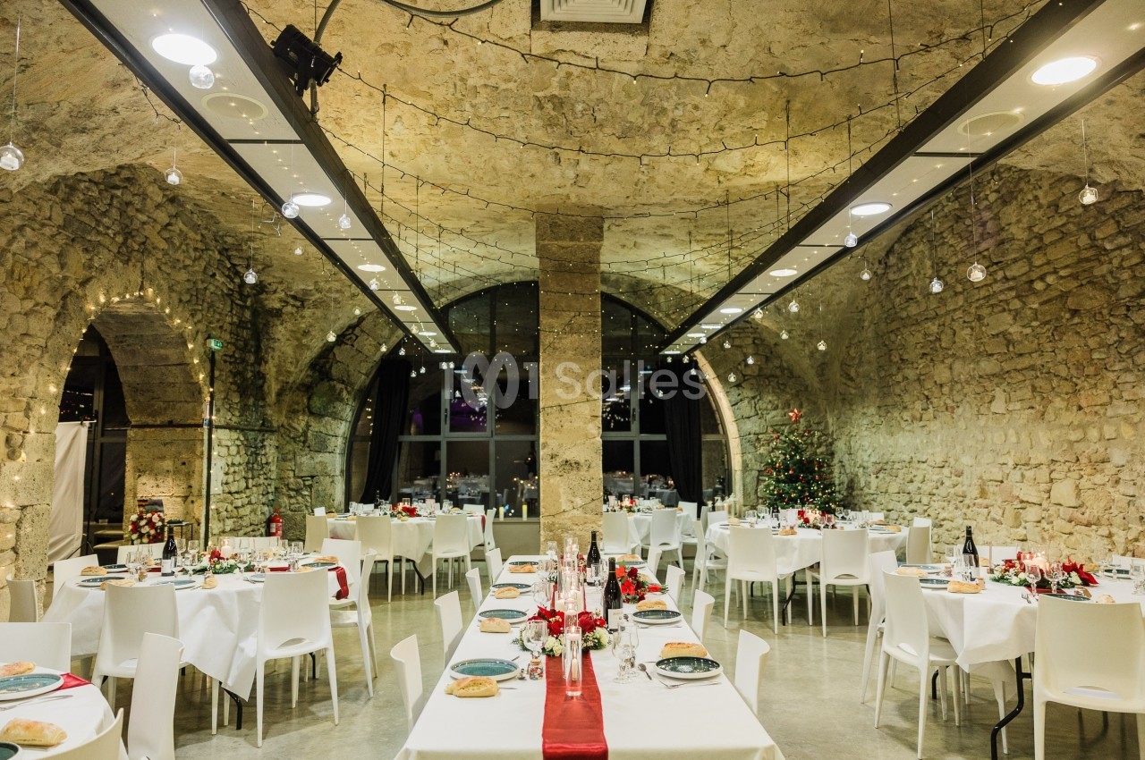 Salle en pierre voûtée décorée pour un repas festif, avec tables dressées, nappes blanches et guirlandes lumineuses.