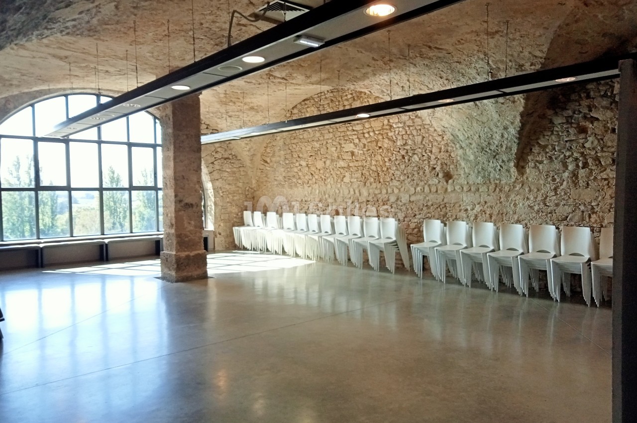 Salle lumineuse avec murs en pierre, grandes fenêtres cintrées et rangée de chaises blanches alignées contre un mur.