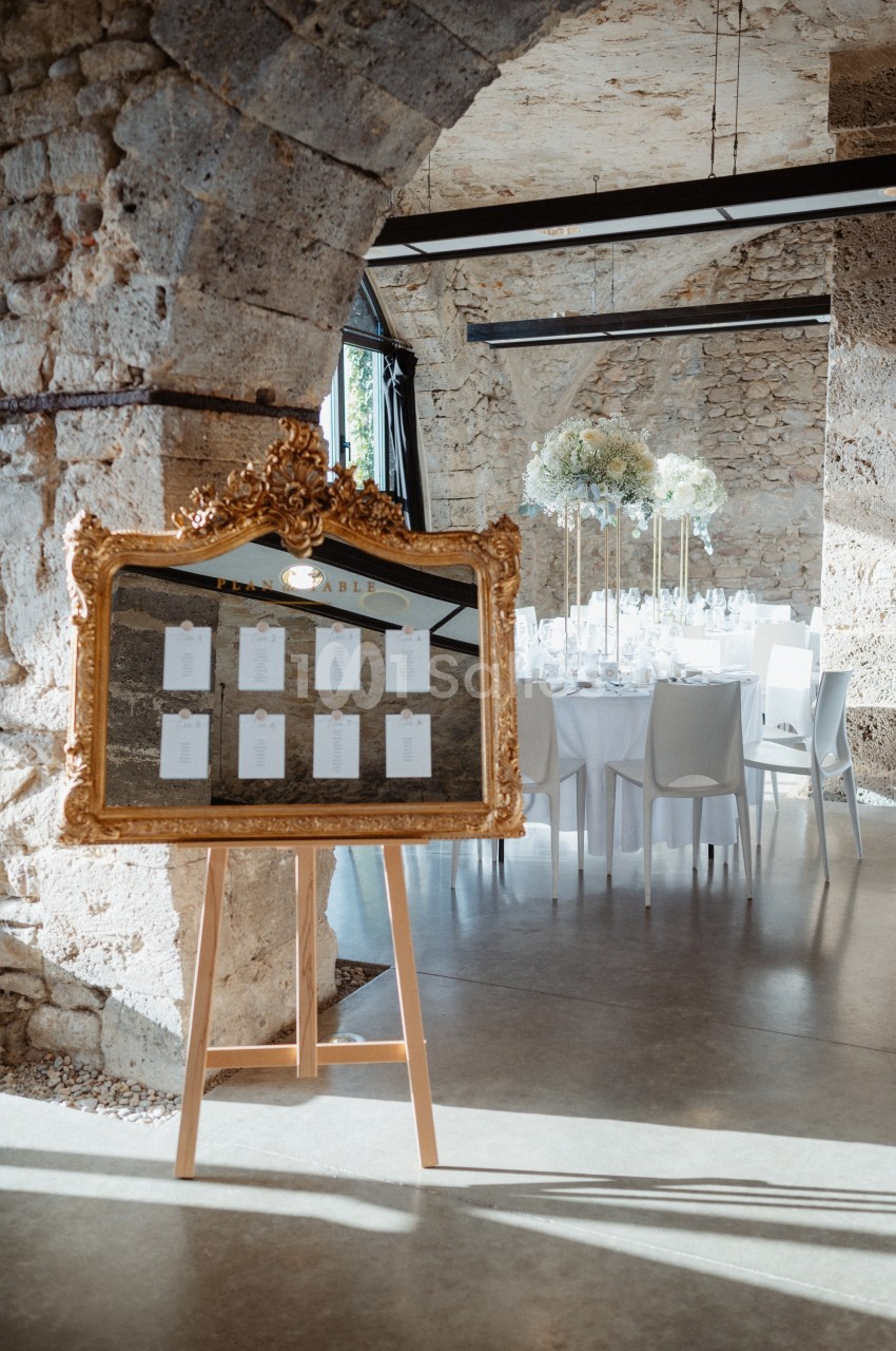 Plan de table encadré en or sur un chevalet, devant une salle de réception décorée avec des tables et des fleurs blanches.