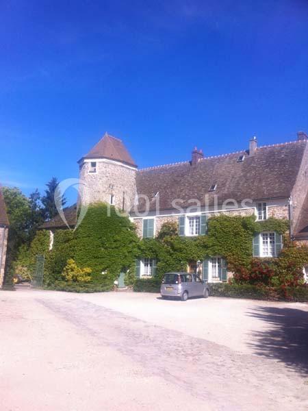 Grande maison en pierre avec tour, couverte de végétation, située dans une cour ensoleillée avec une voiture garée.