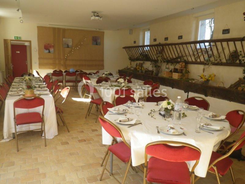 Salle décorée avec des tables rondes dressées, nappes blanches, chaises rouges et éléments rustiques en arrière-plan.