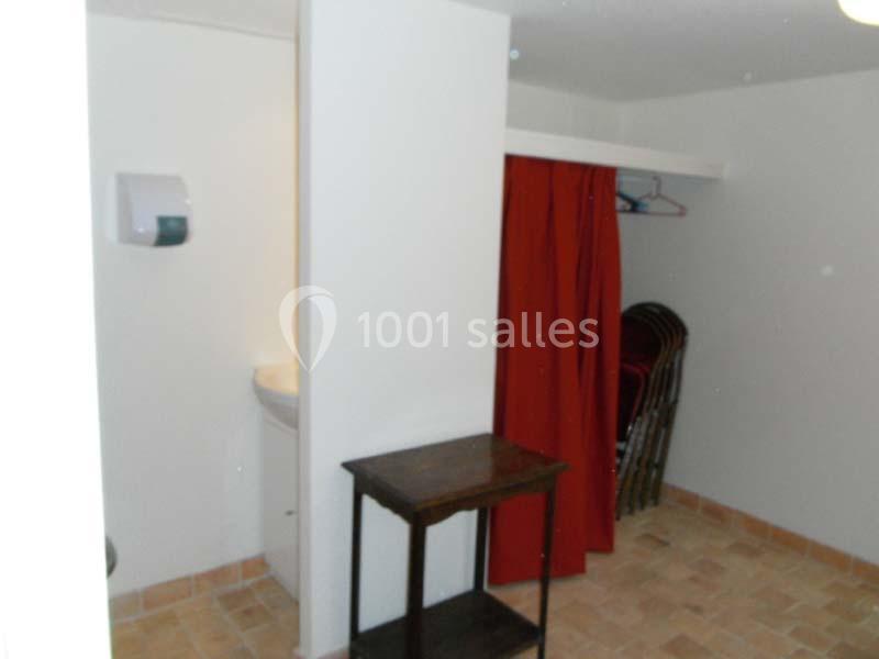 Espace avec un petit lavabo, un distributeur mural, une table en bois et des chaises pliées derrière un rideau rouge.