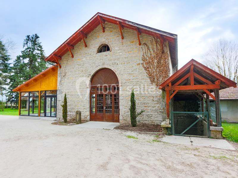Bâtiment en pierre avec façade vitrée, toit en bois et abri adjacent, entouré d'arbres et d'un sol en gravier.