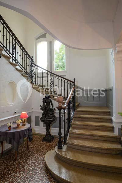 Escalier intérieur en bois avec rampe en fer forgé, éclairé par une grande fenêtre, table et lampe à gauche.