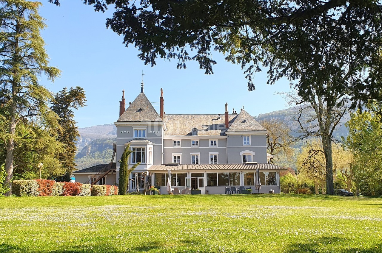 Manoir entouré d'un grand jardin verdoyant, avec des montagnes en arrière-plan sous un ciel clair.