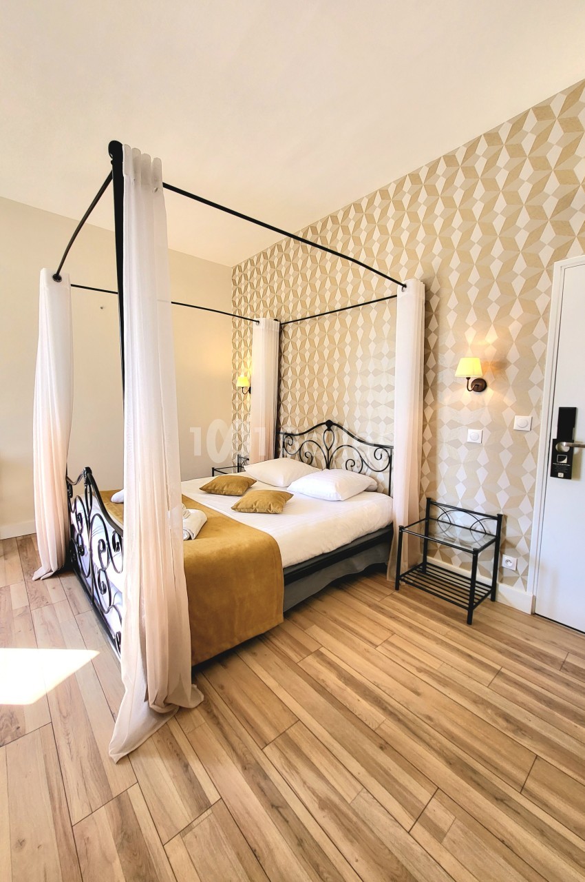 Lit à baldaquin avec voilages blancs dans une chambre lumineuse au sol en bois et murs décorés de motifs géométriques.