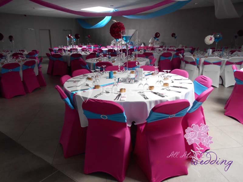 Salle de réception décorée avec des tables rondes dressées, nappes blanches et chaises roses ornées de rubans bleus.