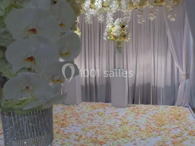Décoration florale avec des orchidées blanches et des pétales au sol, devant des rideaux clairs et un vase central.