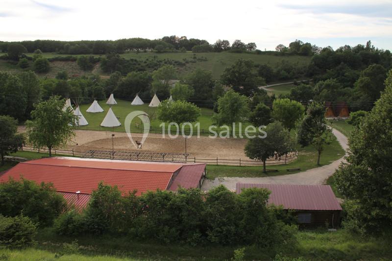 Vue d'un terrain avec des tipis blancs, une zone sablonneuse avec des animaux et des bâtiments entourés de verdure.