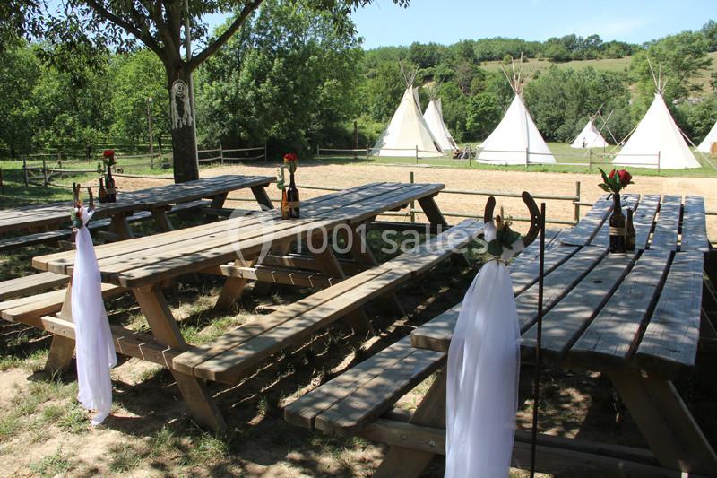 Tables de pique-nique en bois décorées de fleurs et de tissus blancs, avec des tipis en arrière-plan dans un cadre naturel.
