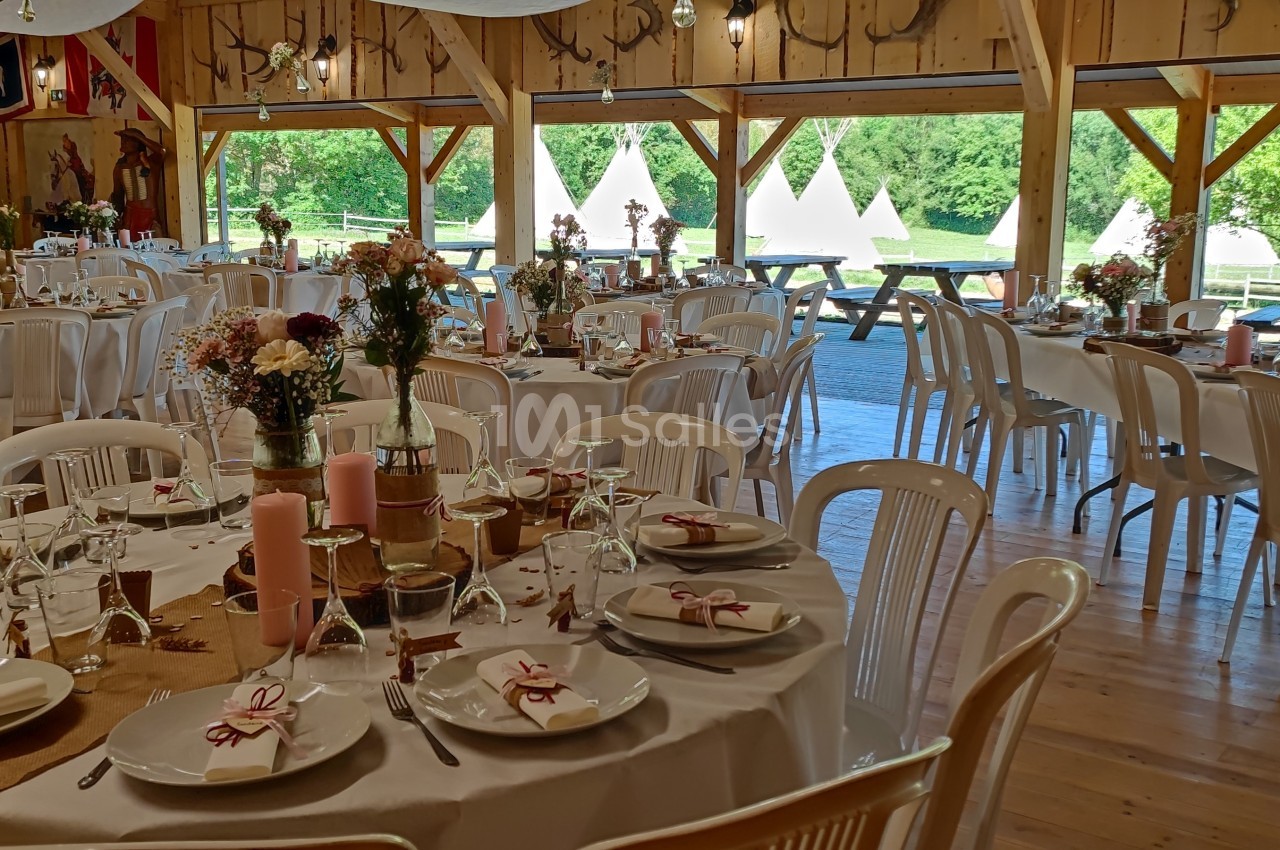 Salle décorée pour un événement avec tables rondes dressées, chaises blanches et vue sur un extérieur verdoyant avec tipis.