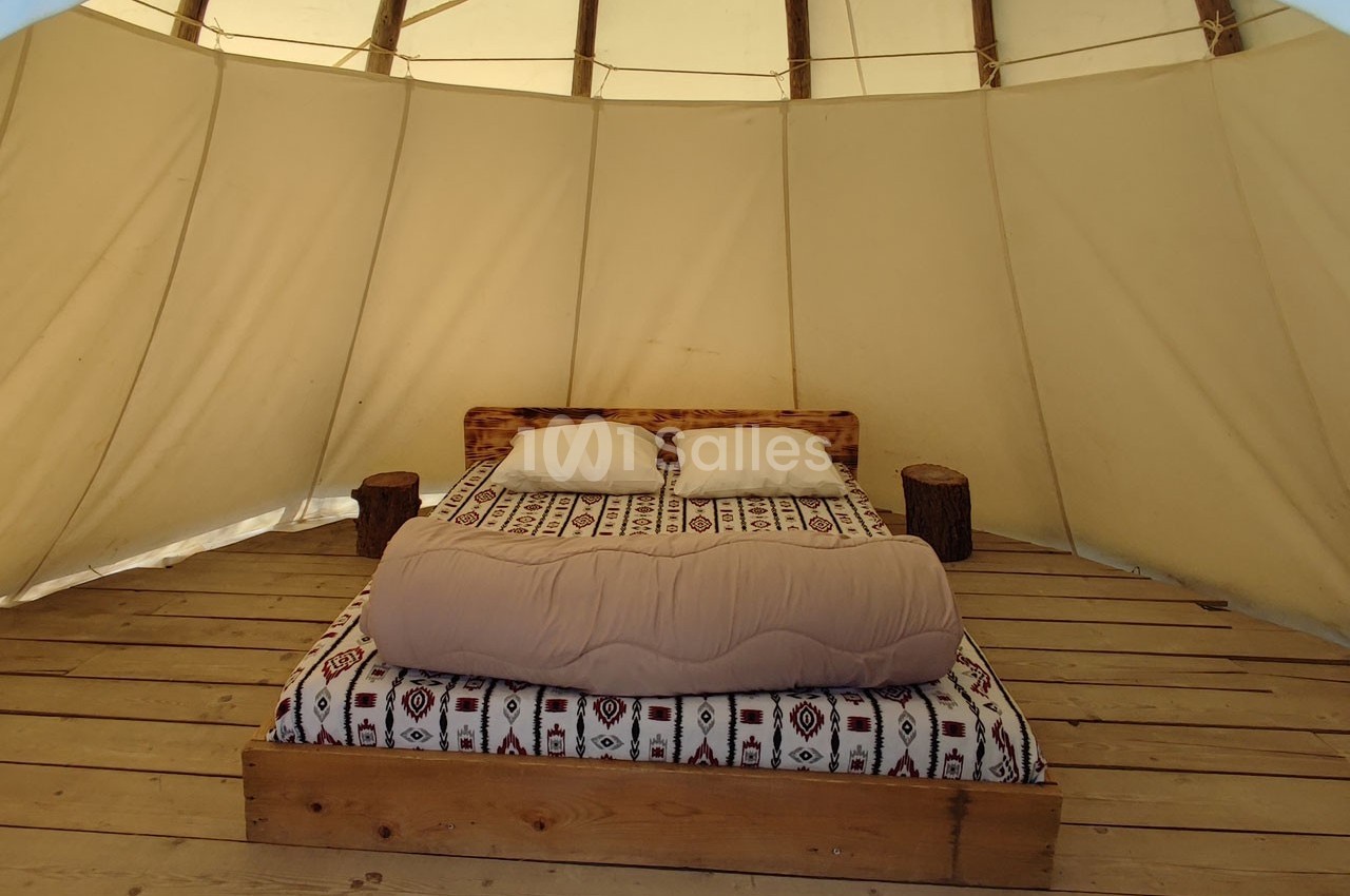 Lit double avec couverture beige et coussins blancs, installé dans une tente avec structure en bois et toile beige.
