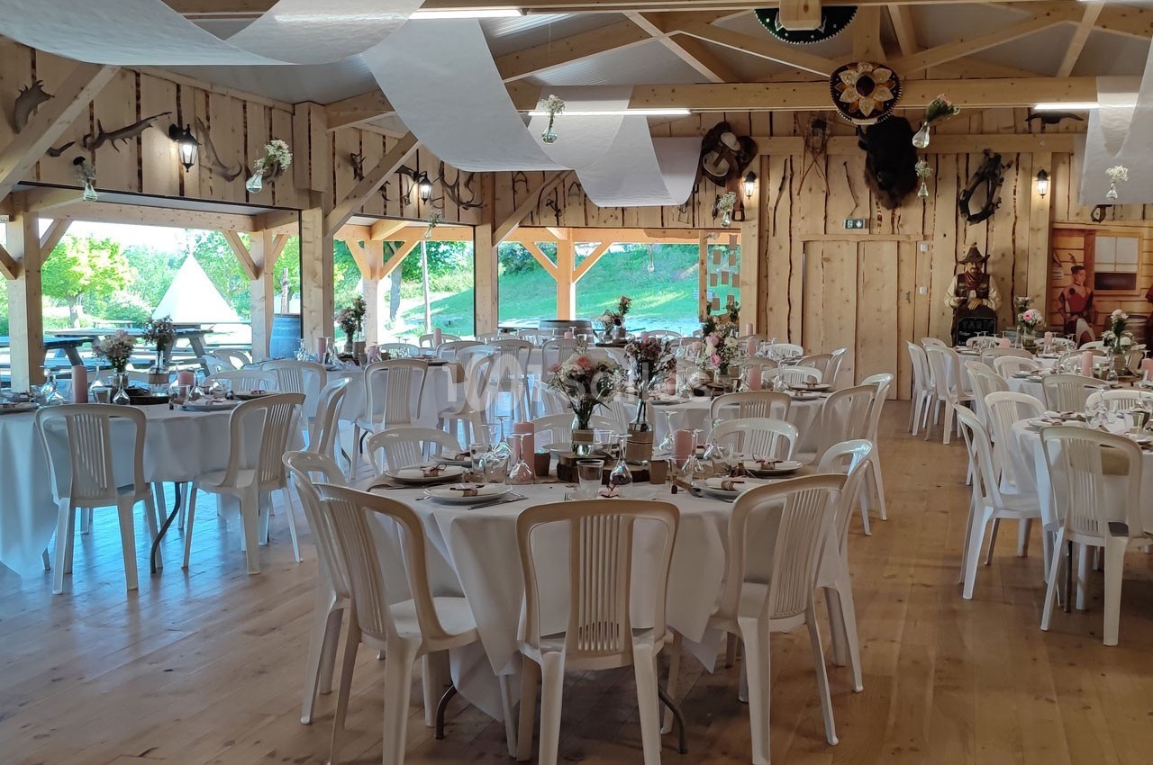 Salle de réception en bois décorée pour un événement, avec tables rondes dressées et chaises blanches.