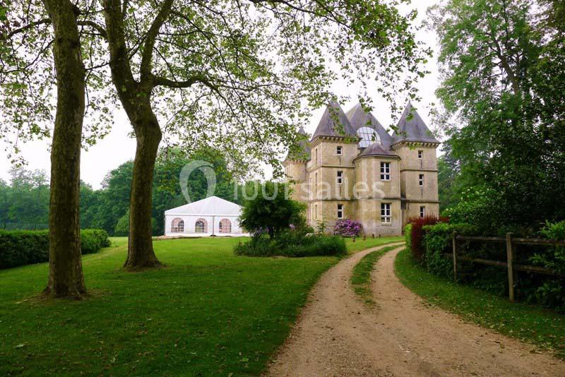 Allée gravillonnée menant à un château entouré d'arbres, avec une tente blanche installée sur la pelouse.