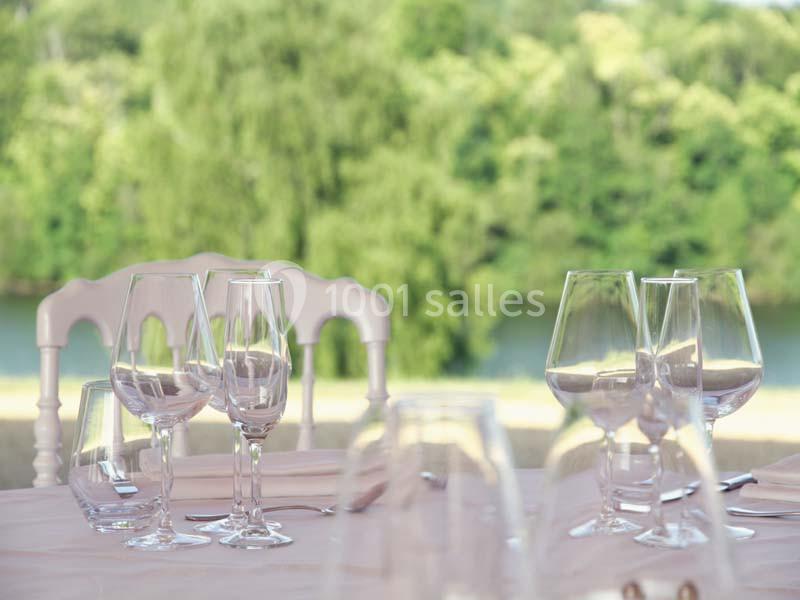 Table dressée avec des verres à pied, vue floue sur un paysage verdoyant et une rivière en arrière-plan.