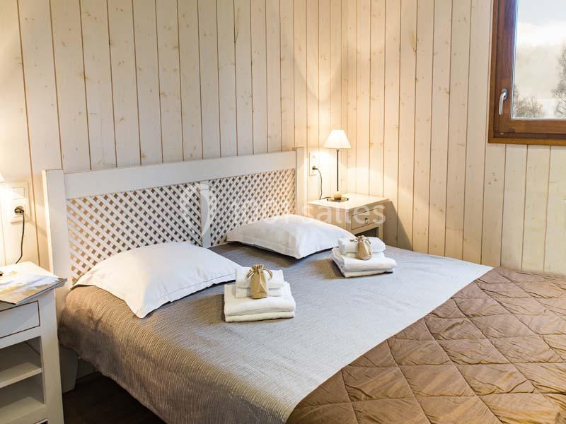 Chambre lumineuse avec lit double, draps blancs, plaid marron, tables de chevet et mur en bois clair.