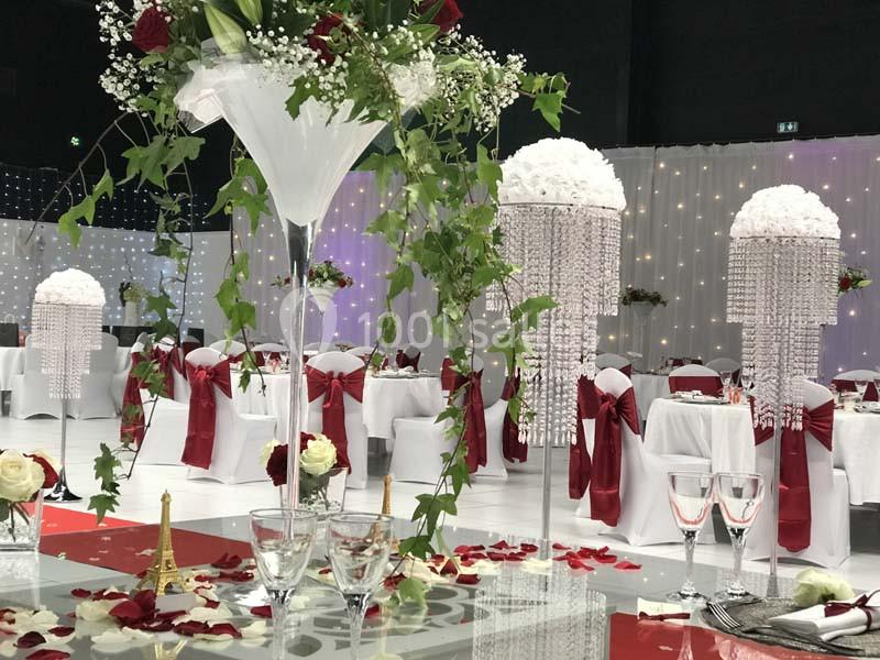 Salle de réception décorée avec des tables ornées de roses rouges, nappes blanches et chaises habillées de rubans rouges.