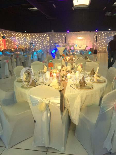 Salle de réception décorée avec des tables rondes, nappes blanches, chaises habillées et guirlandes lumineuses en arrière…