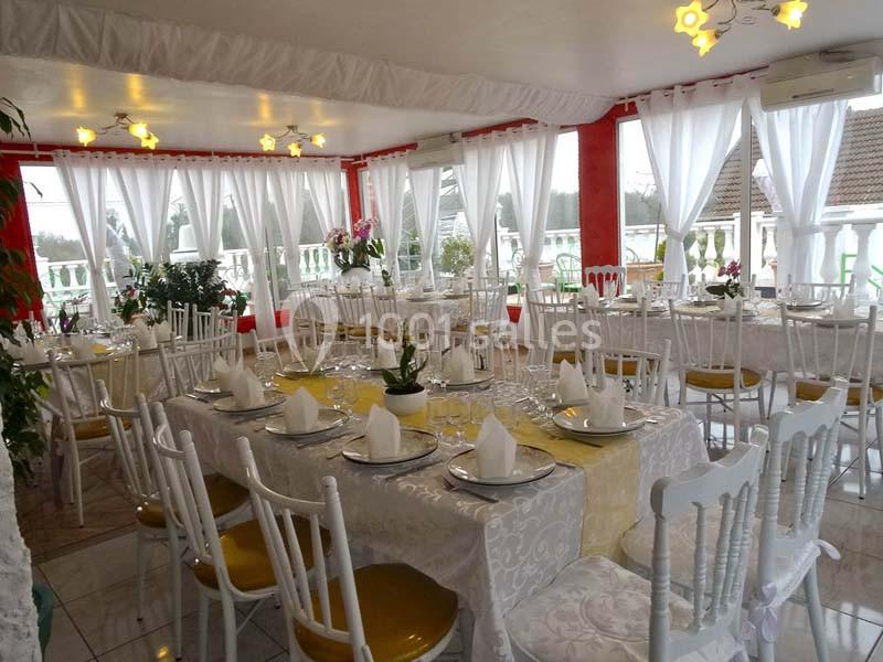 Salle de réception lumineuse avec tables décorées de nappes blanches, vaisselle élégante et chaises blanches.