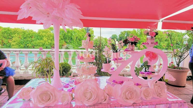 Table décorée en rose avec fleurs, gâteaux et ornements sous une tente, vue sur un jardin et une rivière en arrière-plan.