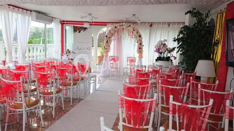 Salle décorée pour une cérémonie avec des chaises blanches ornées de rubans rouges et une arche florale en arrière-plan.