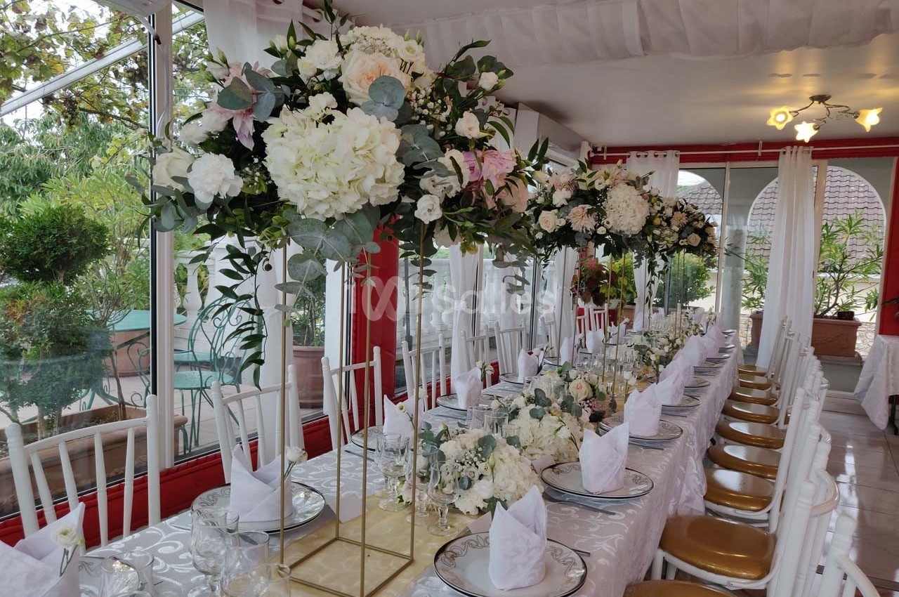 Table décorée pour un événement avec nappes blanches, chaises dorées et arrangements floraux élégants sur supports hauts.
