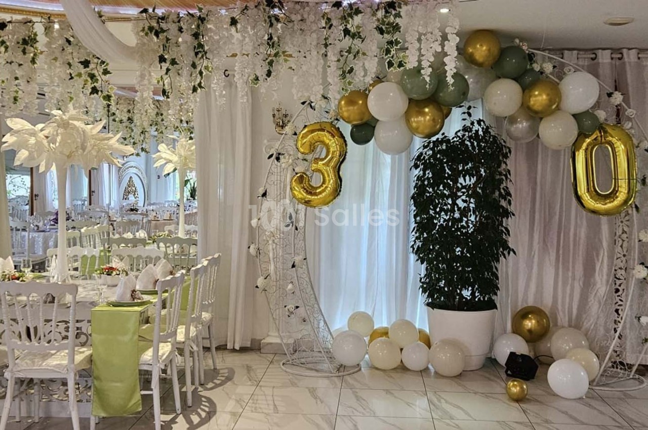 Décoration de salle de fête avec arche de ballons dorés et blancs formant le chiffre 30, plantes et tables dressées.