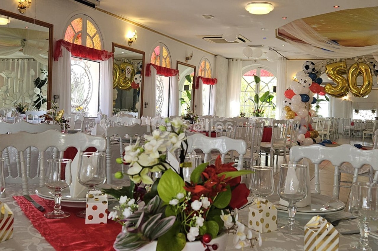 Salle de réception décorée avec des tables dressées, des ballons colorés et une arche marquée ’50’.