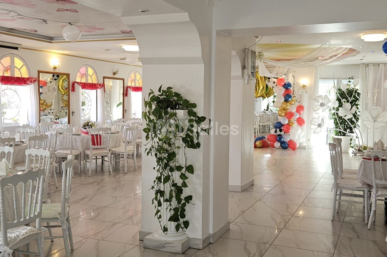 Salle de réception lumineuse décorée avec des tables dressées, des plantes et des ballons colorés au fond.