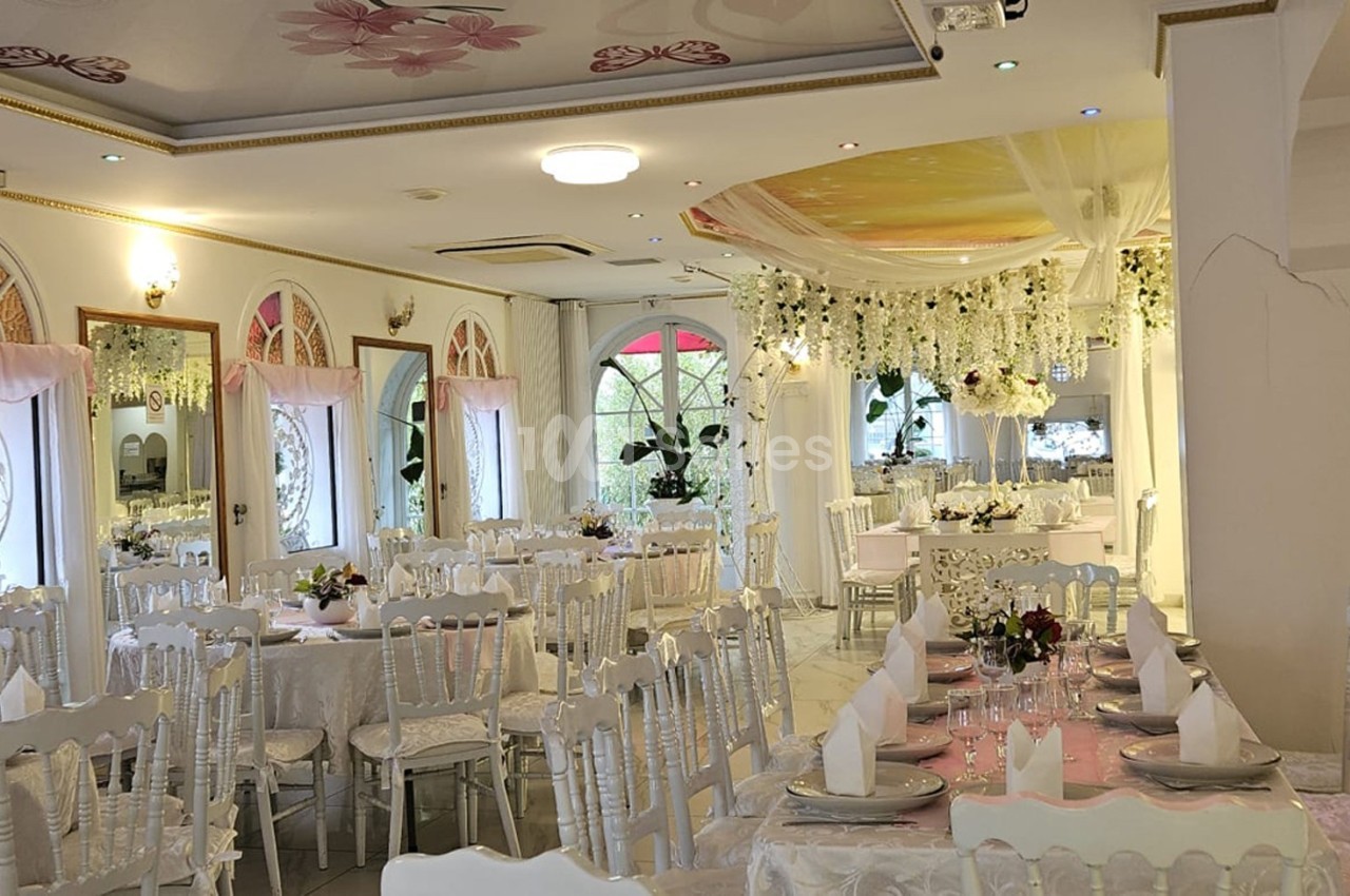 Salle de réception lumineuse décorée de fleurs blanches, tables dressées avec nappes blanches et chaises élégantes.