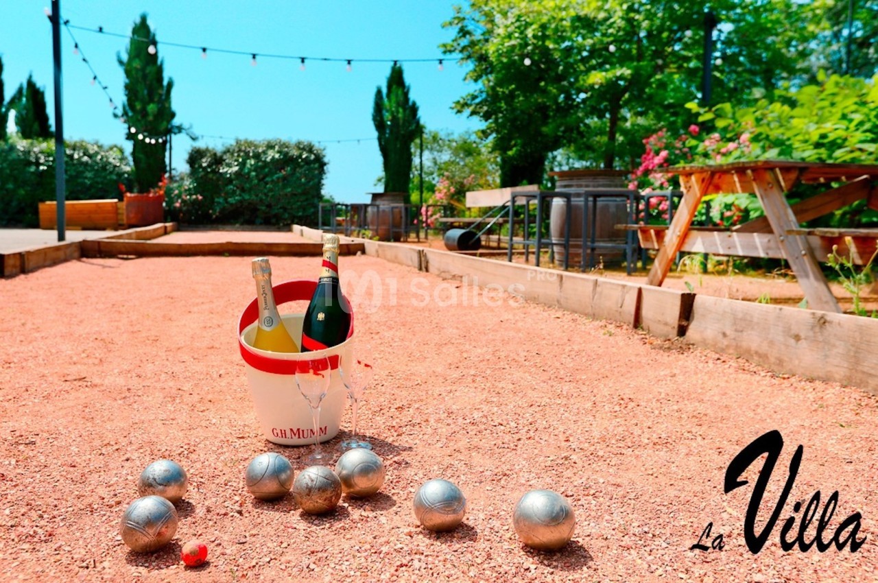 Terrain de pétanque avec des boules en métal, un seau contenant des bouteilles et des tables en bois à l'arrière-plan.