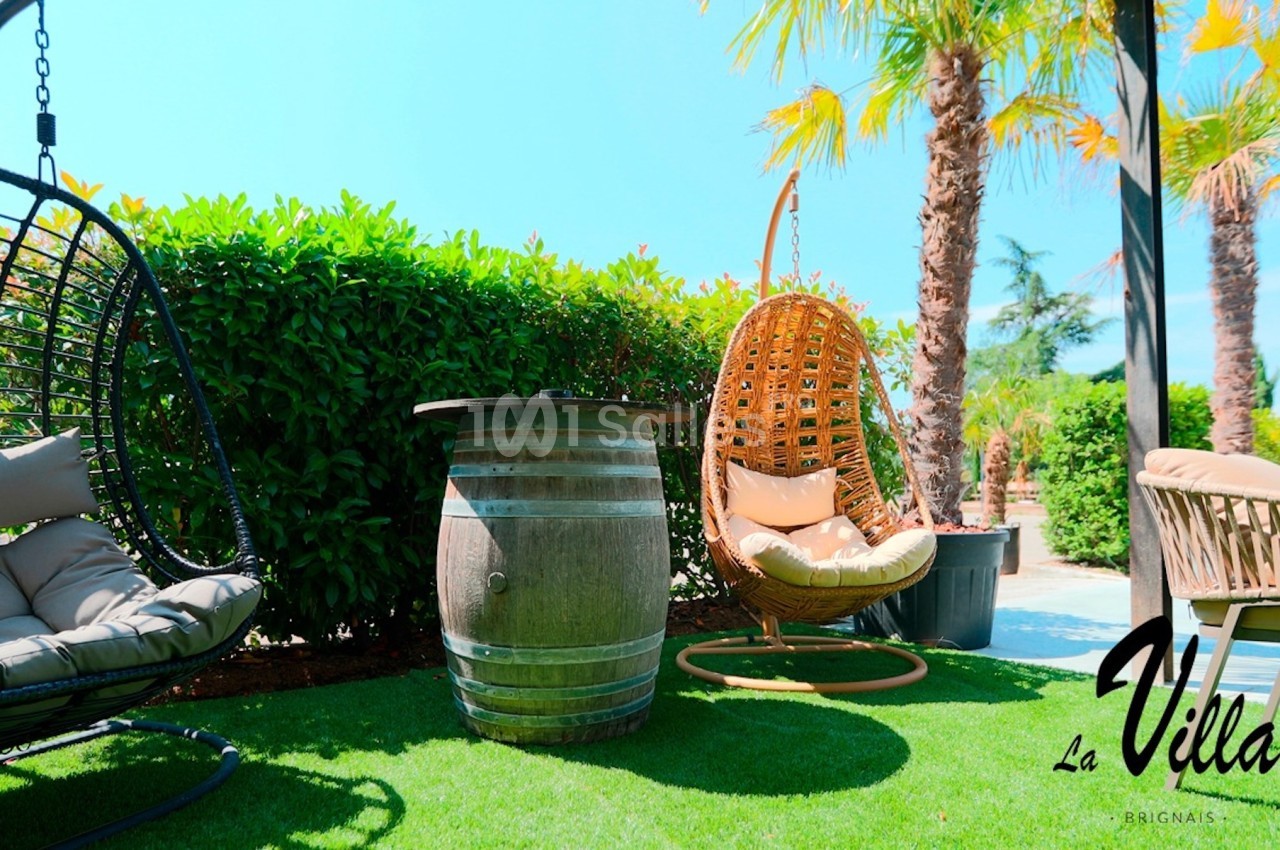 Mobilier de jardin avec fauteuils suspendus, tonneau en bois en guise de table, sur pelouse entourée de palmiers.