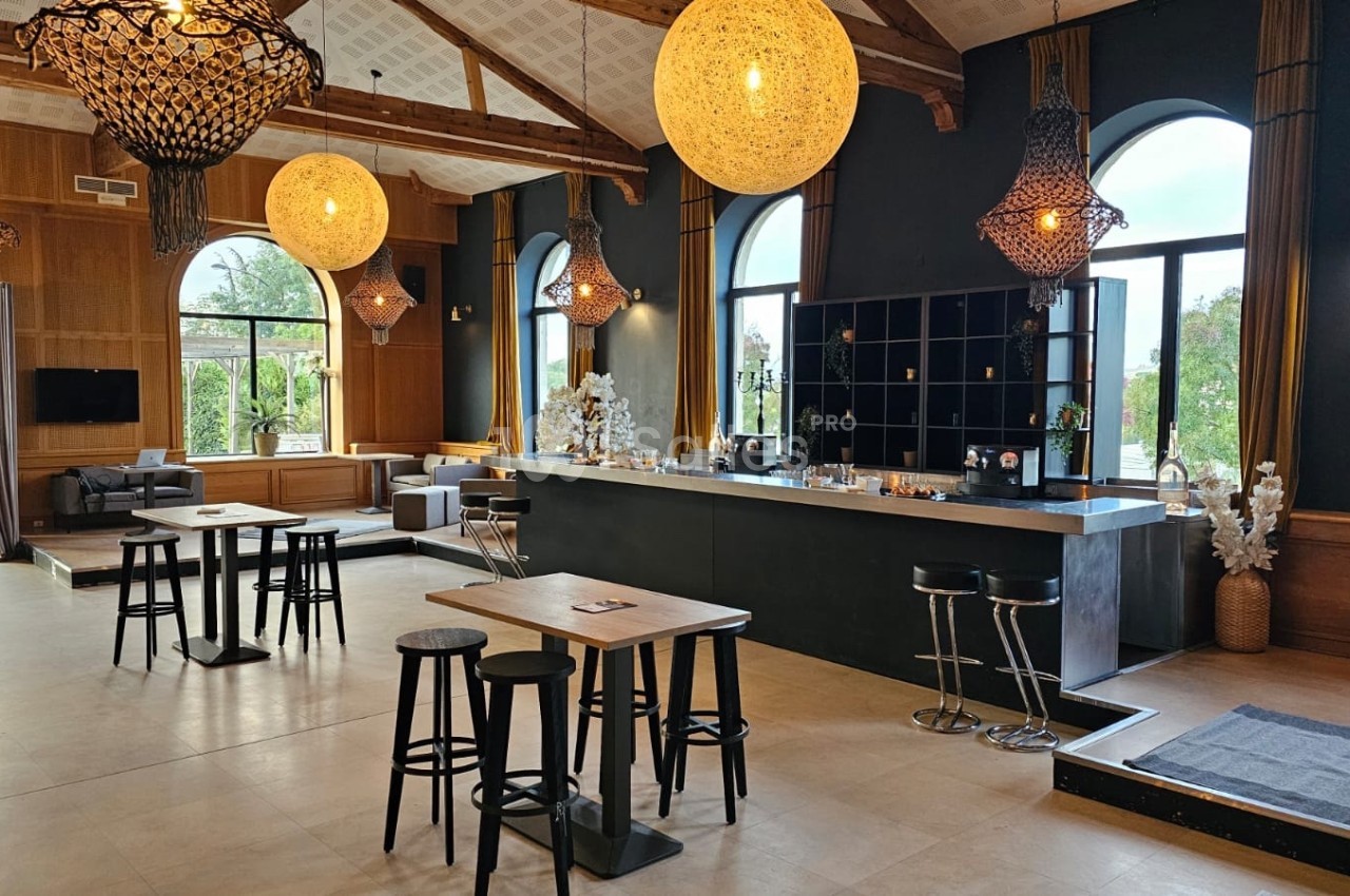Salle lumineuse avec un bar central, tables hautes en bois, grandes fenêtres cintrées et décorations modernes.