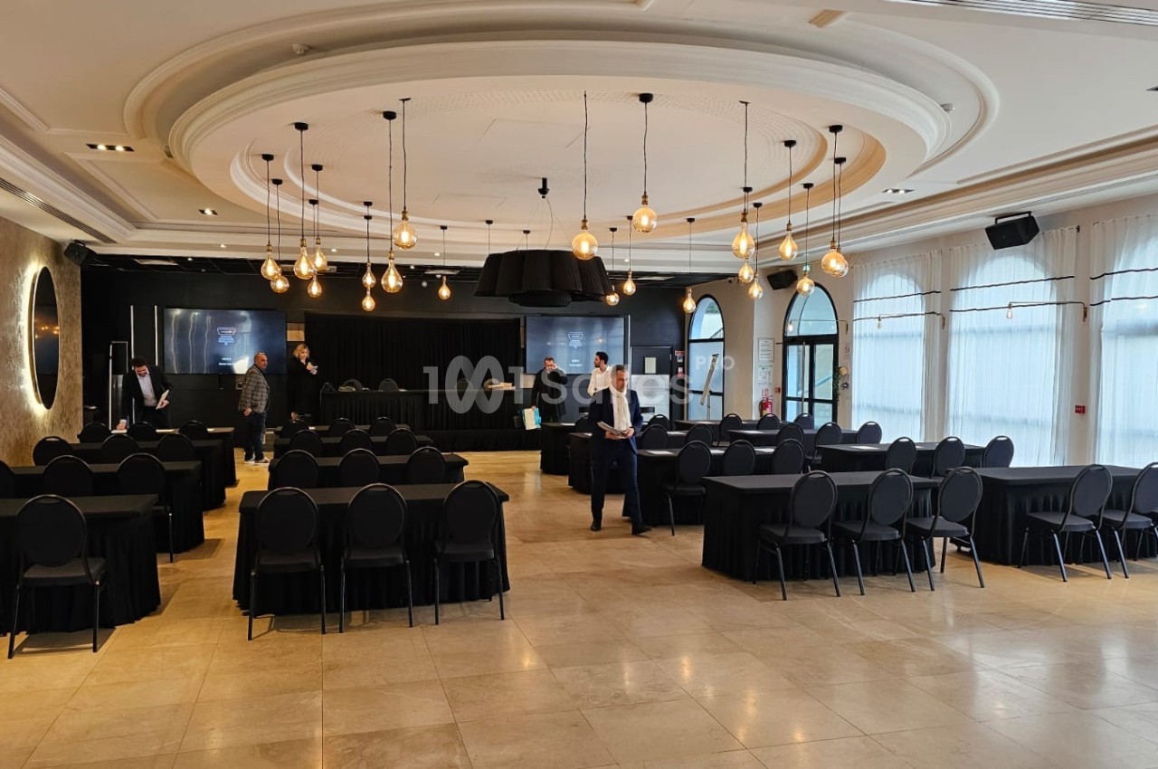 Salle de conférence lumineuse avec tables et chaises noires, préparée pour un événement, quelques personnes présentes.
