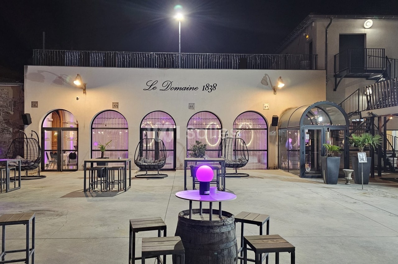 Cour extérieure éclairée la nuit avec tables, chaises, lampes violettes et façade d'un bâtiment ’Le Domaine 1838’.