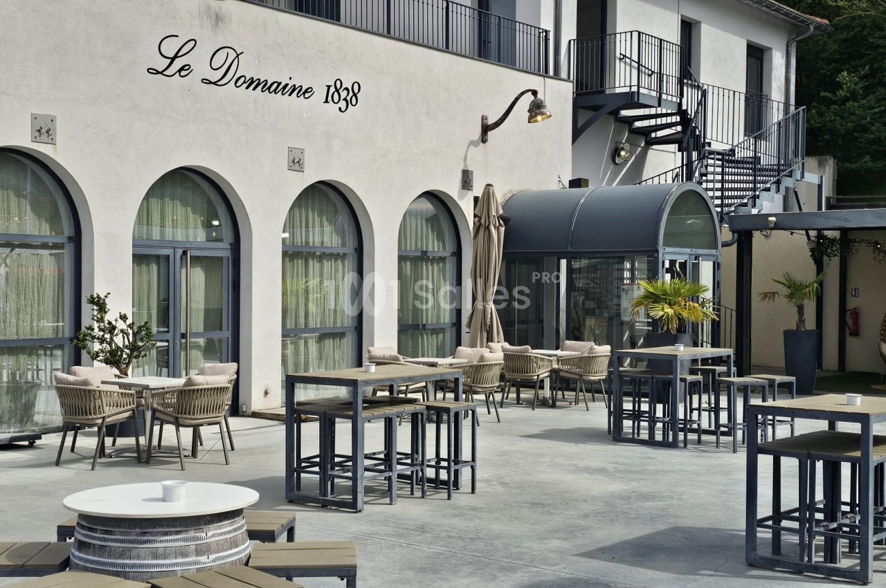 Terrasse extérieure avec tables, chaises et parasols devant un bâtiment aux grandes fenêtres et escaliers métalliques.