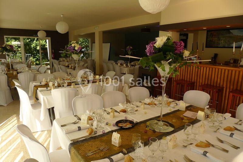 Salle de réception décorée avec des tables dressées, nappes blanches, centres de table fleuris et lumière naturelle.
