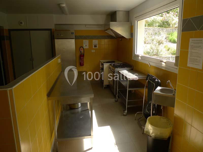 Cuisine professionnelle avec murs jaunes, plan de travail en inox, évier, cuisinière, fenêtre et équipements de rangement.
