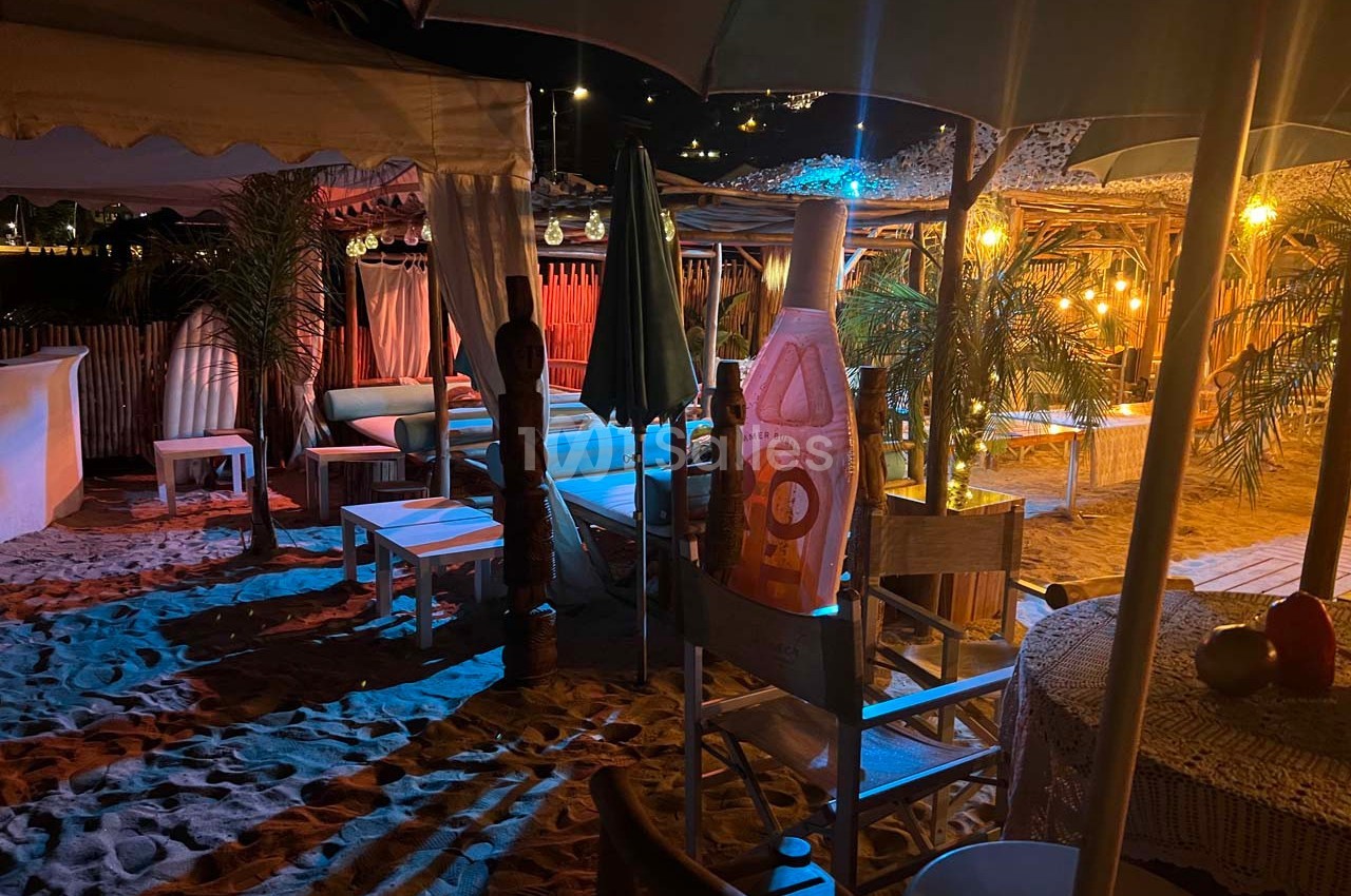 Terrasse de plage éclairée la nuit, avec chaises, tables, parasols et décorations en bois sous des lumières tamisées.