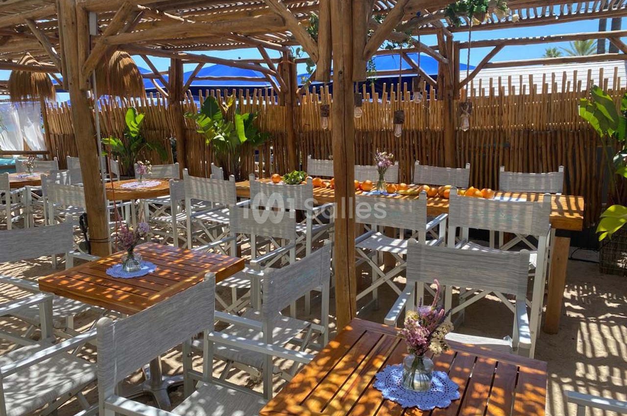 Terrasse en bois ombragée avec tables et chaises blanches, décorée de fleurs et entourée de palissades en bambou.
