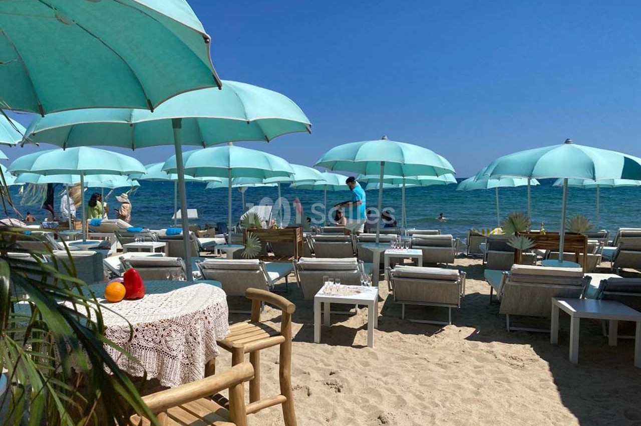 Plage aménagée avec des parasols turquoise, des transats et des tables sur le sable, face à la mer calme.