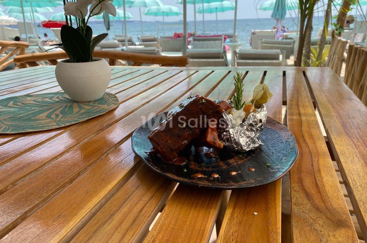 Assiette de côtes grillées avec garniture, servie sur une table en bois face à une plage avec parasols et transats.