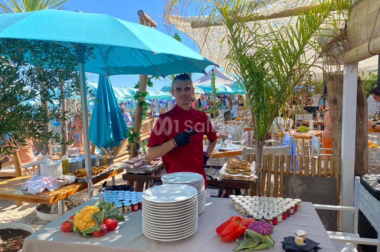 Un serveur devant une table dressée avec des assiettes, légumes et condiments, dans un restaurant en plein air.
