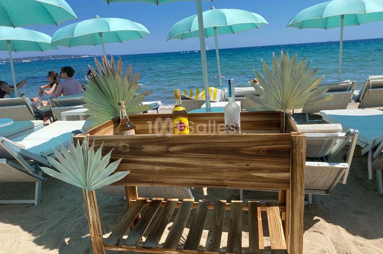 Chariot en bois avec bouteilles, placé sur une plage sous des parasols turquoise près de transats face à la mer.