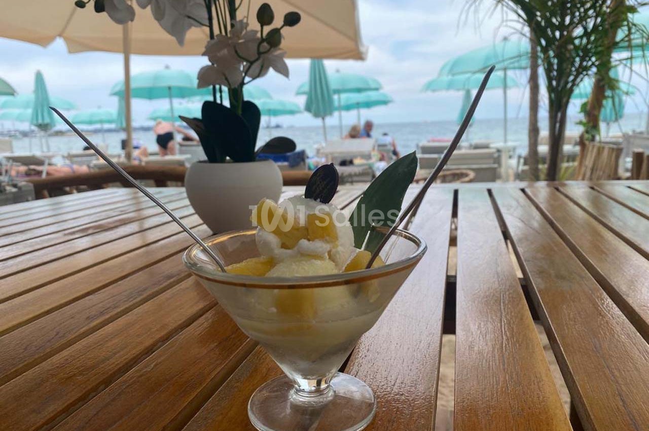 Coupe de glace avec morceaux d'ananas, servie sur une table en bois en terrasse face à la mer avec parasols verts.