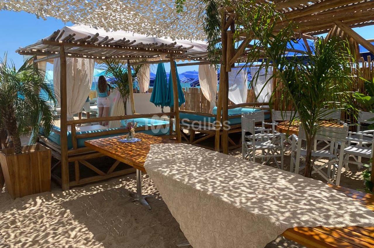 Espace de détente sur une plage avec lits baldaquins, table en bois, chaises et ombrage partiel sous un filet.