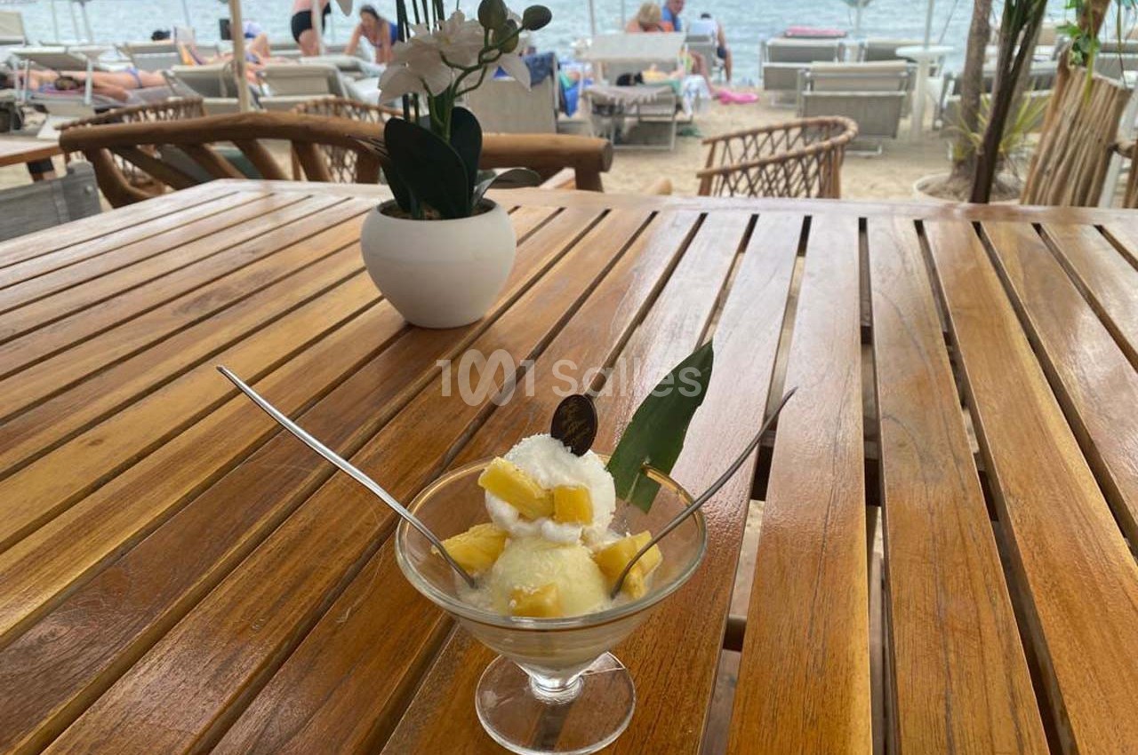 Coupe de glace garnie de morceaux de mangue et d'une feuille, posée sur une table en bois face à une plage.