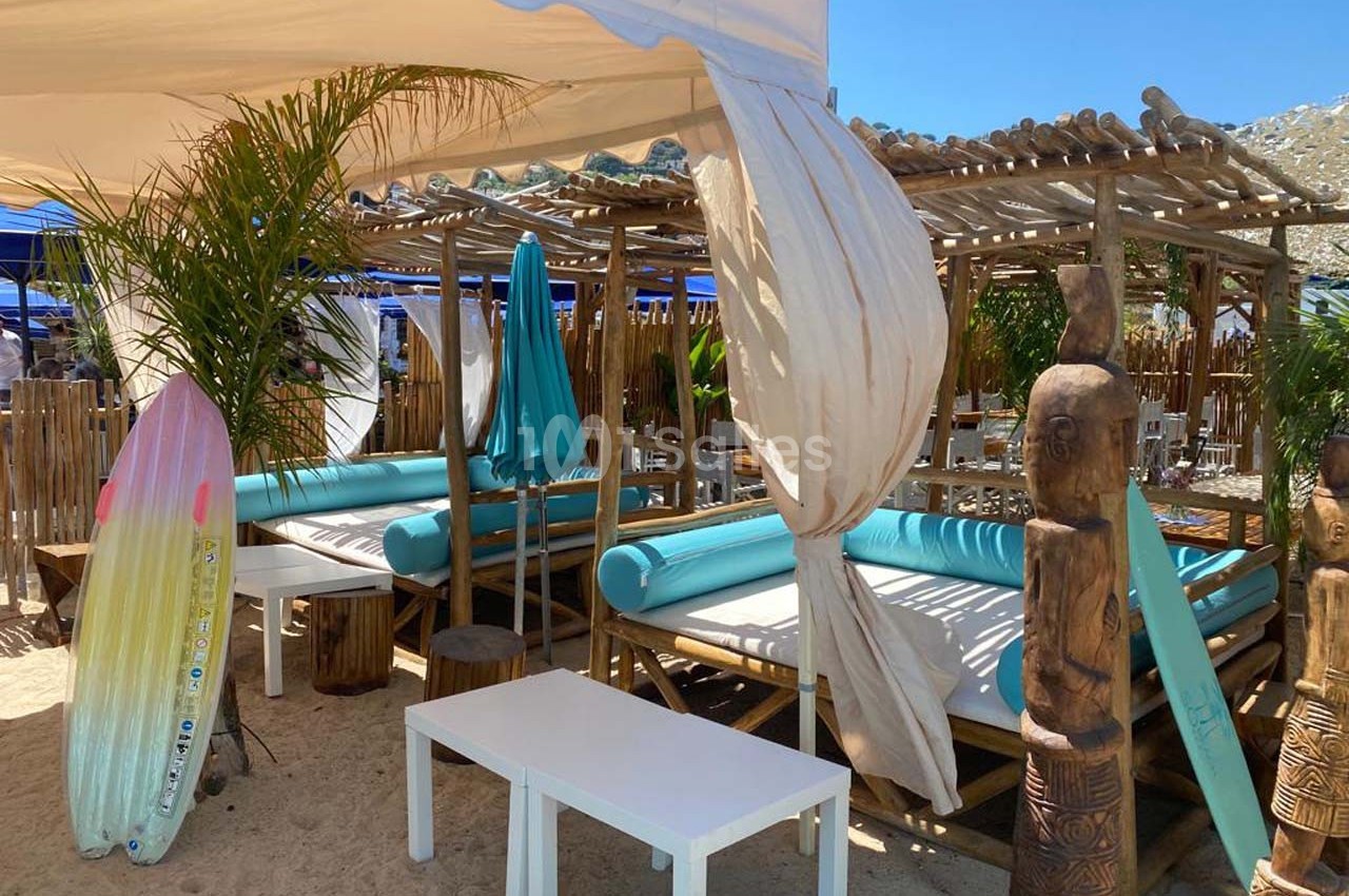 Terrasse en bois avec lits de plage turquoise, parasols et décorations tropicales, sur du sable en plein soleil.